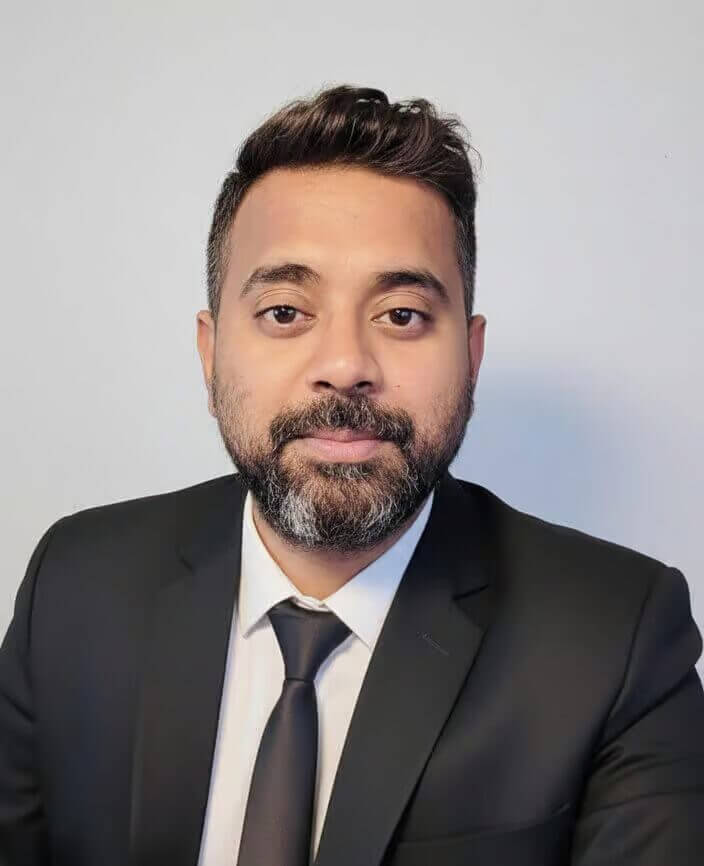 Professional headshot of a man in a black suit and tie, with a neutral background.